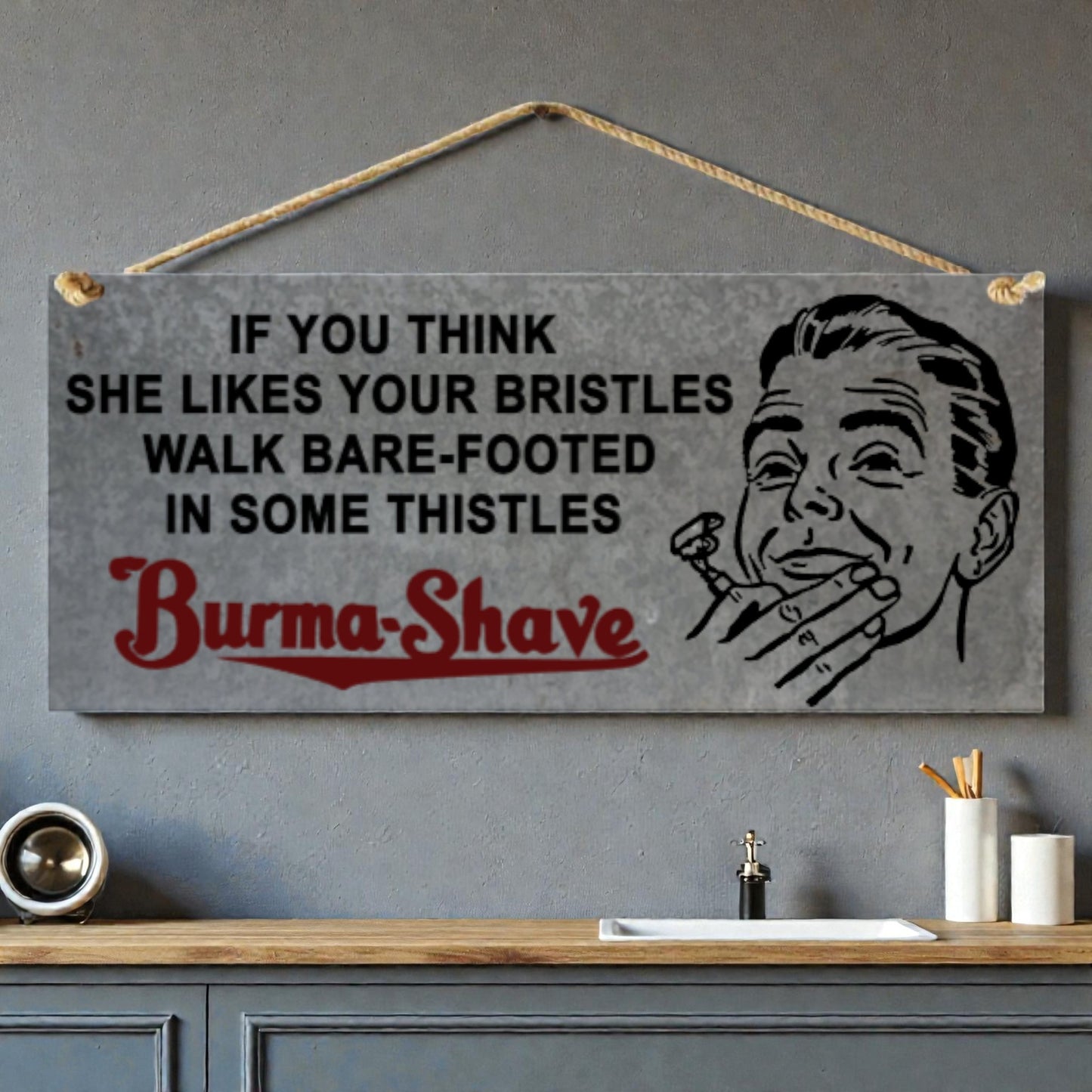 Vintage-style sign with humorous text and illustration of a man holding a razor, hanging on a wall above a bathroom counter.