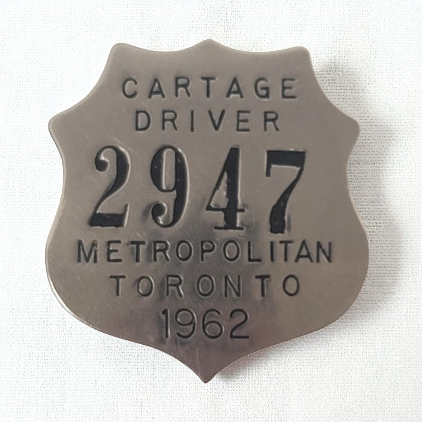 Metallic badge with text 'CARTAGE DRIVER 2947 METROPOLITAN TORONTO 1962' on a white background