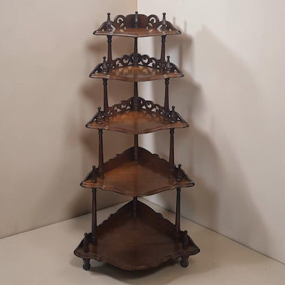 A five-tier wooden corner shelf with ornate scrollwork details and small caster wheels, shown in a studio corner setting.