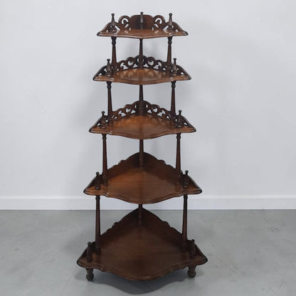 A vintage five-tier wooden corner shelf with ornate scrollwork details and small caster wheels, shown against a plain light gray wall and concrete floor.