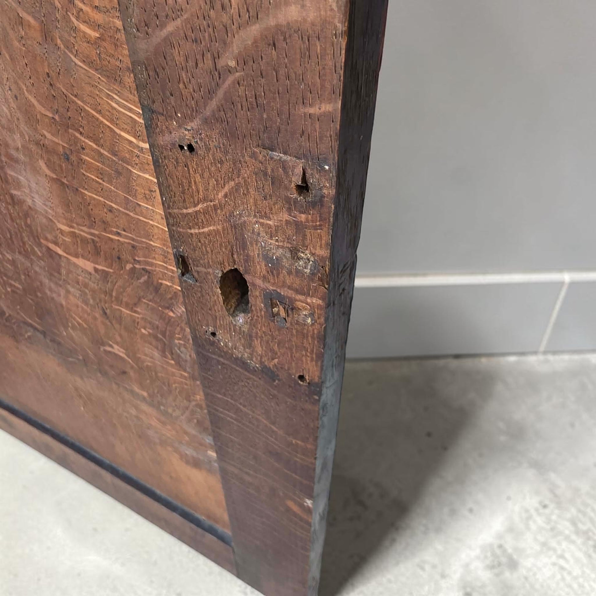 Close-up of a dark brown wooden panel or door showing the wood grain, knots, and edge against a light gray tiled wall and concrete floor.