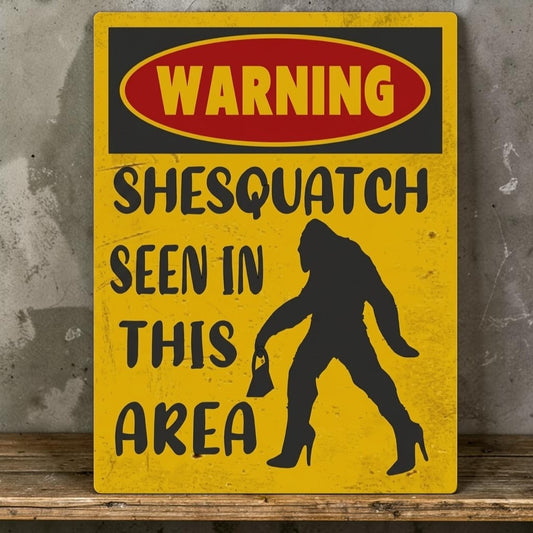 A novelty warning sign with a yellow background, a red oval header reading 'WARNING', black text saying 'SHESQUATCH SEEN IN THIS AREA', and a black silhouette of a Bigfoot-like figure wearing high heels, shown leaning against a gray wall on a wooden surface.