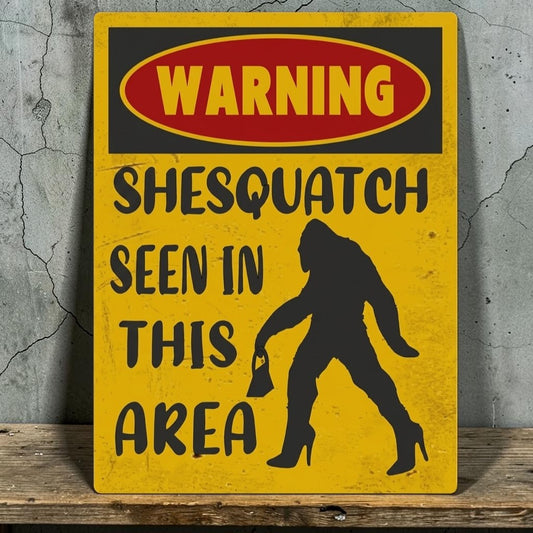A novelty warning sign with a distressed yellow background, a red and black oval header reading 'WARNING', and the text 'SHESQUATCH SEEN IN THIS AREA' alongside a black silhouette of a Bigfoot-like figure in high heels, shown leaning against a gray concrete wall on a wooden surface.