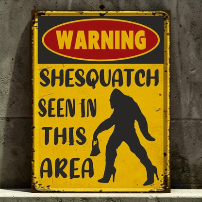 Vintage-style warning sign with a silhouette of a woman and text 'Warning: Shesquatch seen in this area' against a concrete wall.