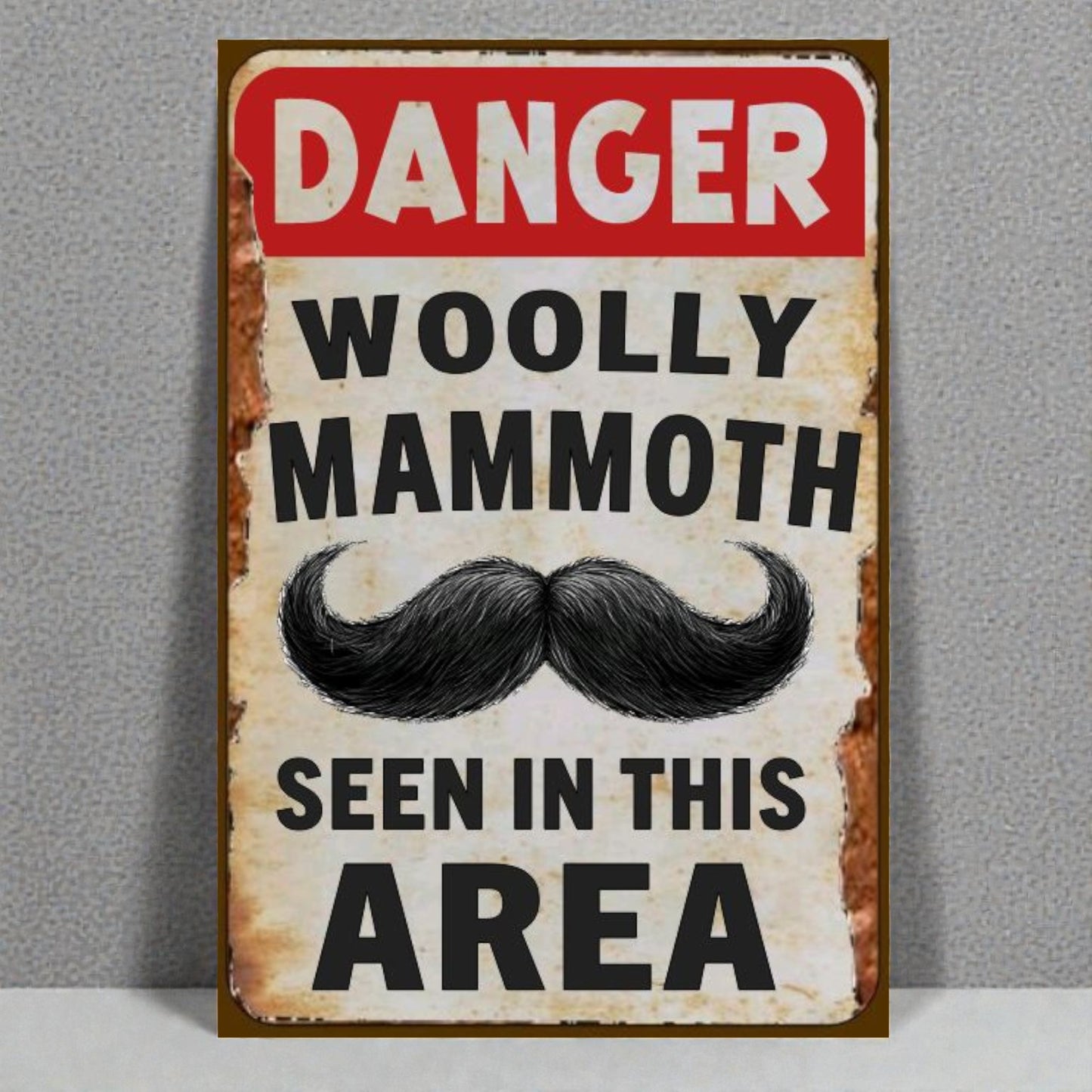 Vintage-style sign with a mustache graphic and text 'Danger Woolly Mammoth Seen in This Area' on a gray background.