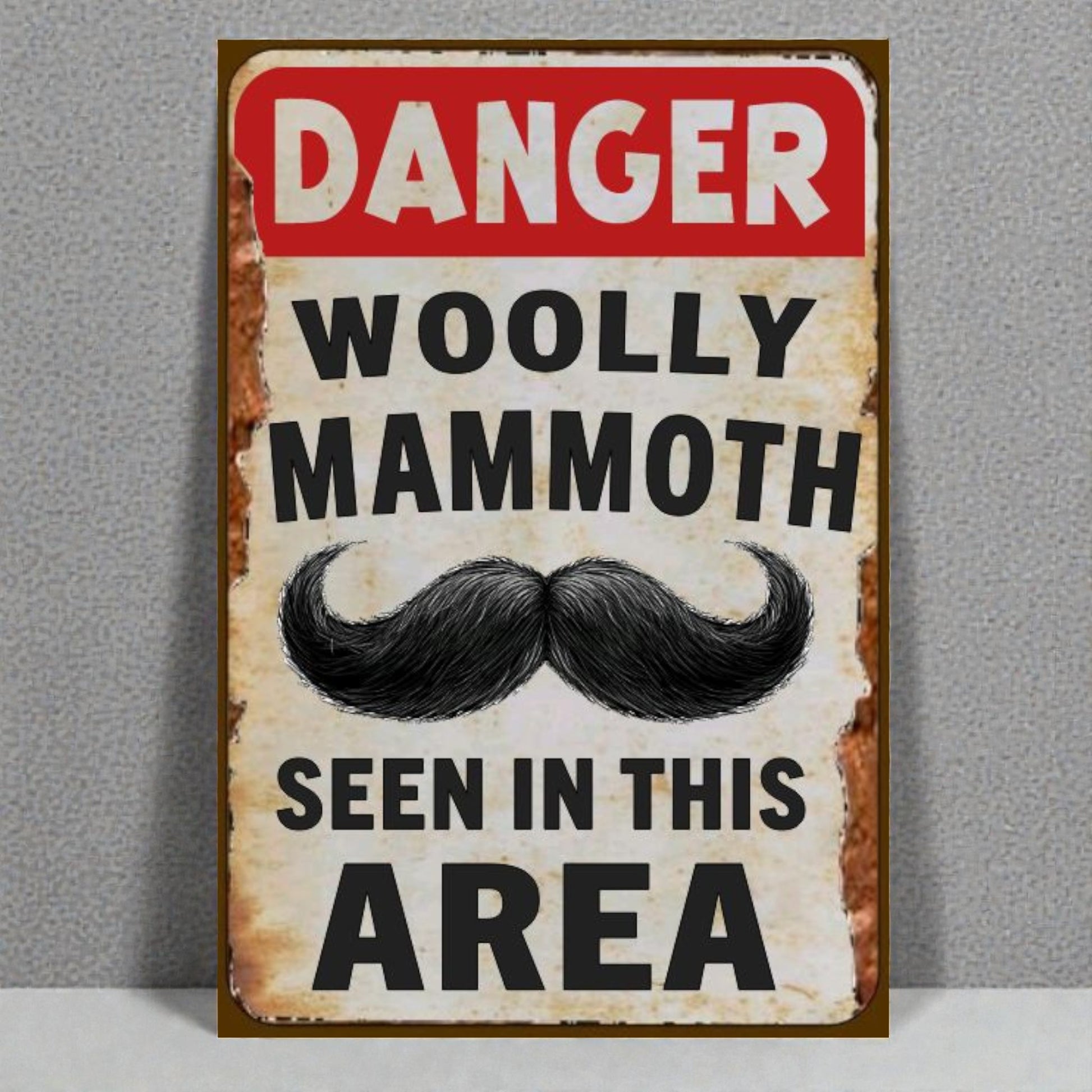 Vintage-style sign with a mustache graphic and text 'Danger Woolly Mammoth Seen in This Area' on a gray background.