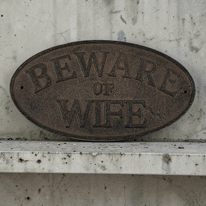 Brown oval sign with 'Beware of Wife' text on a concrete wall.