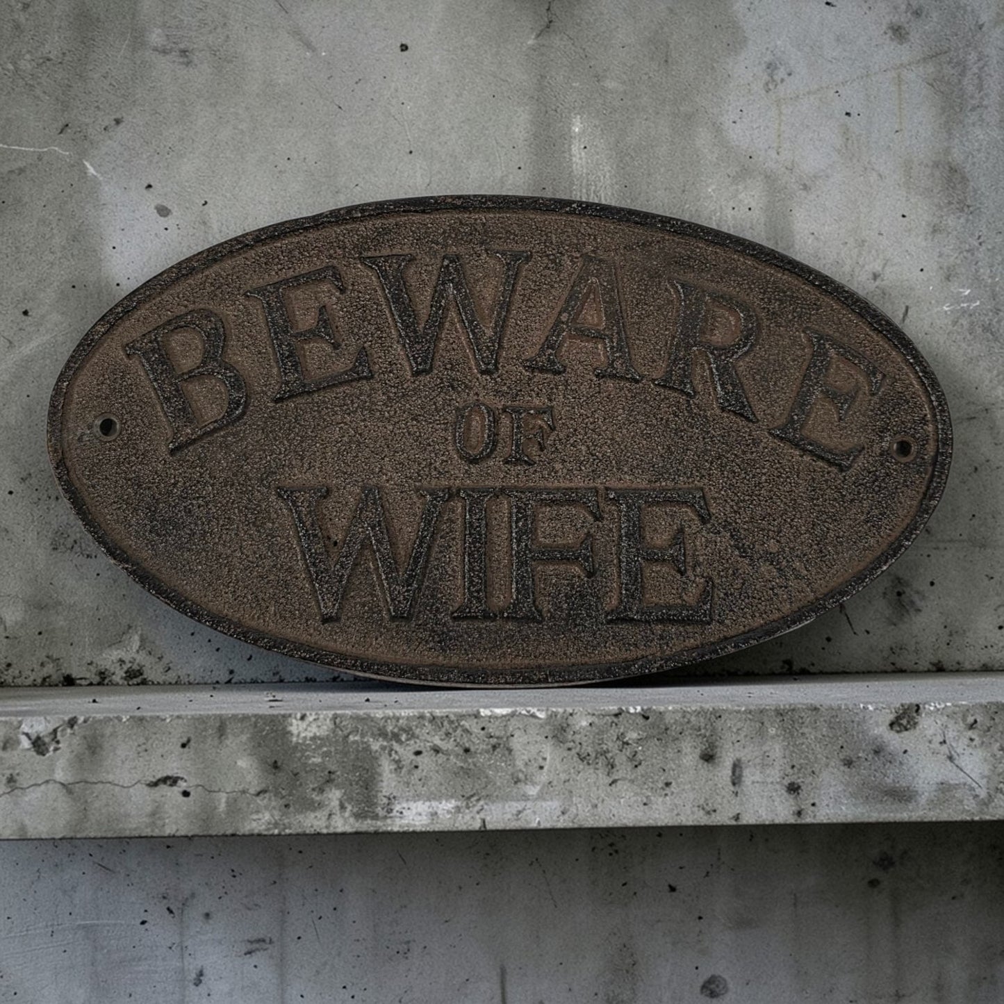 Oval metal sign with 'Beware of Wife' text on a concrete wall.
