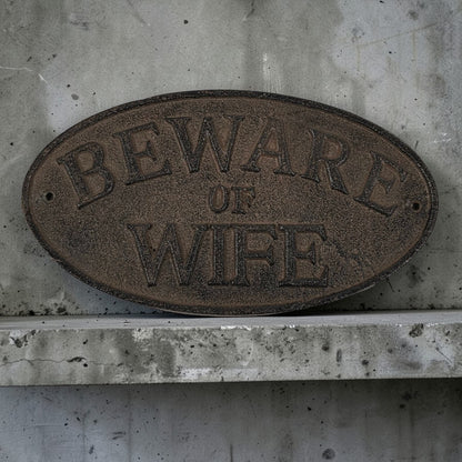 Oval metal sign with 'Beware of Wife' text on a concrete wall.
