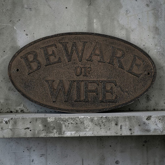 Oval metal sign with 'Beware of Wife' text on a concrete wall.