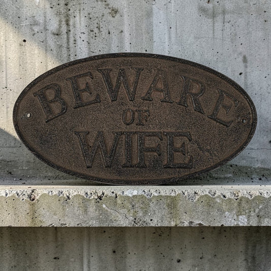 Oval metal sign with 'Beware of Wife' text on a concrete wall.