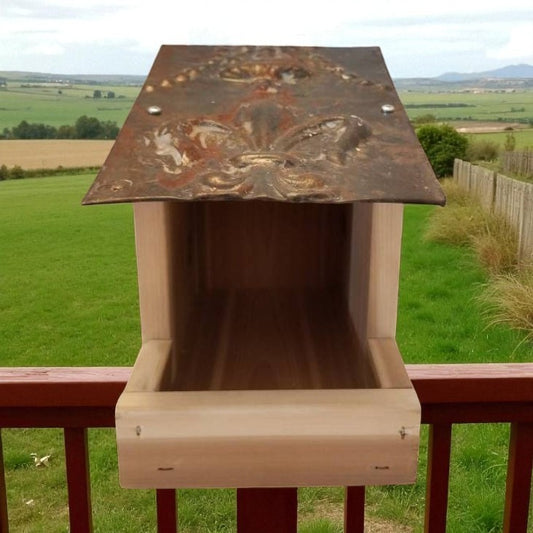 Wooden bird feeder with a metal top on a railing overlooking a green landscape.