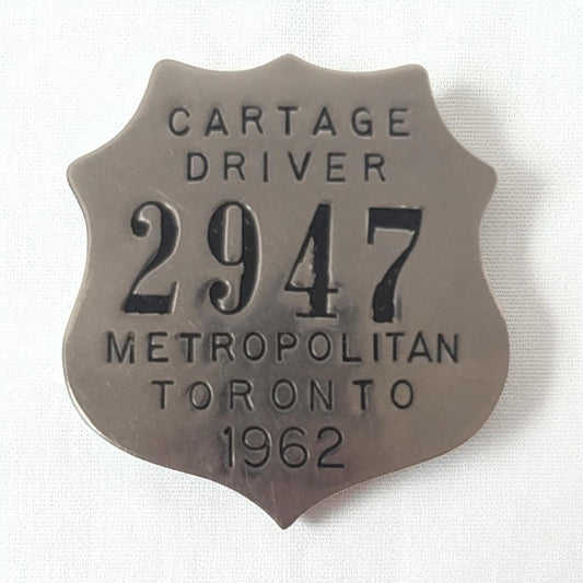 Metallic badge with text 'CARTAGE DRIVER 2947 METROPOLITAN TORONTO 1962' on a white background