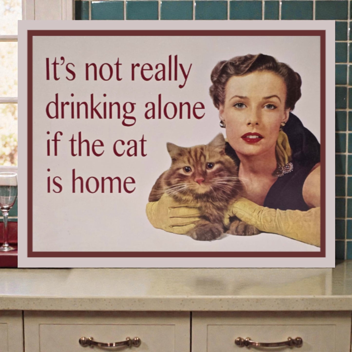 Vintage-style sign with a woman holding a cat and text 'It's not really drinking alone if the cat is home' on a kitchen counter.