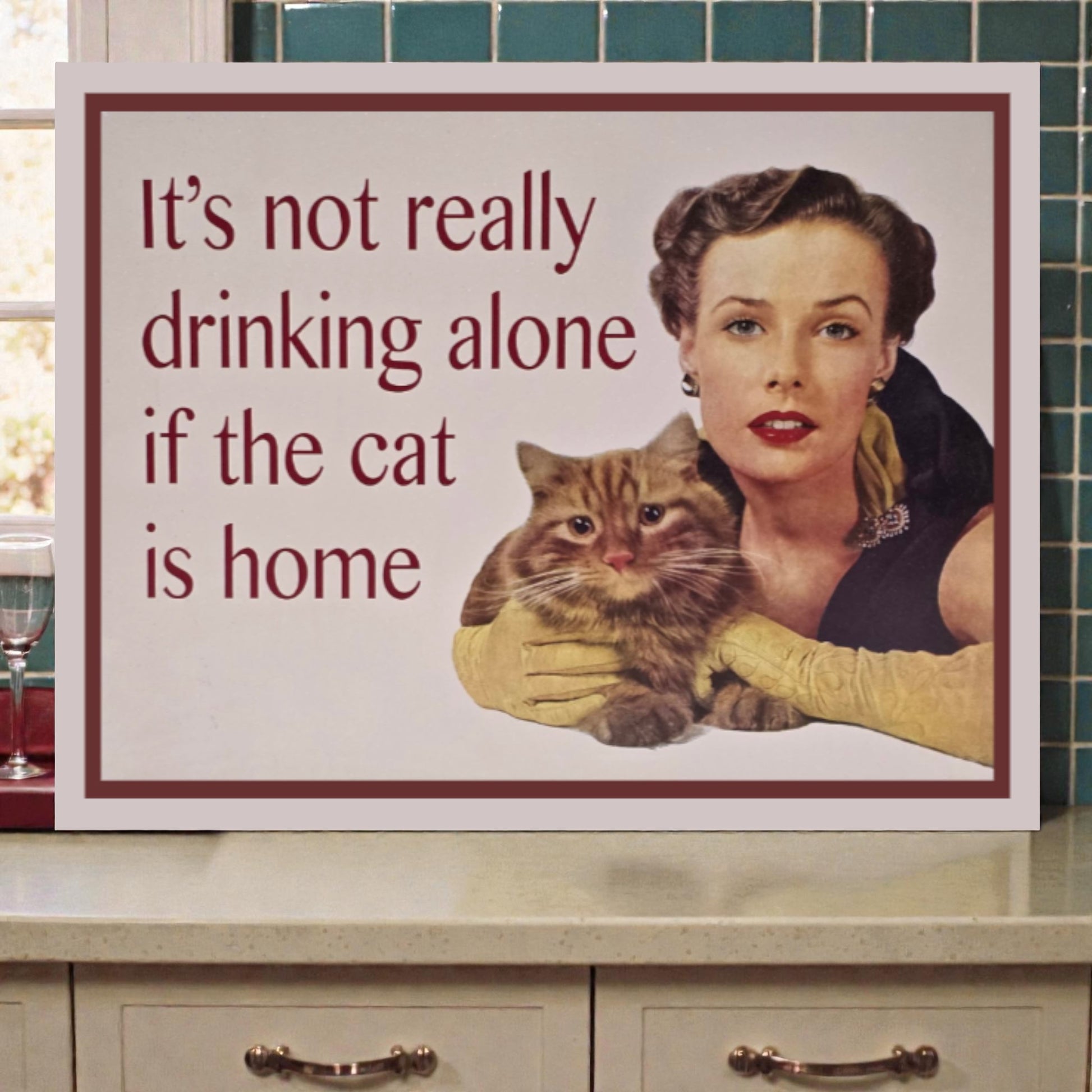 Vintage-style sign with a woman holding a cat and text 'It's not really drinking alone if the cat is home' on a kitchen counter.