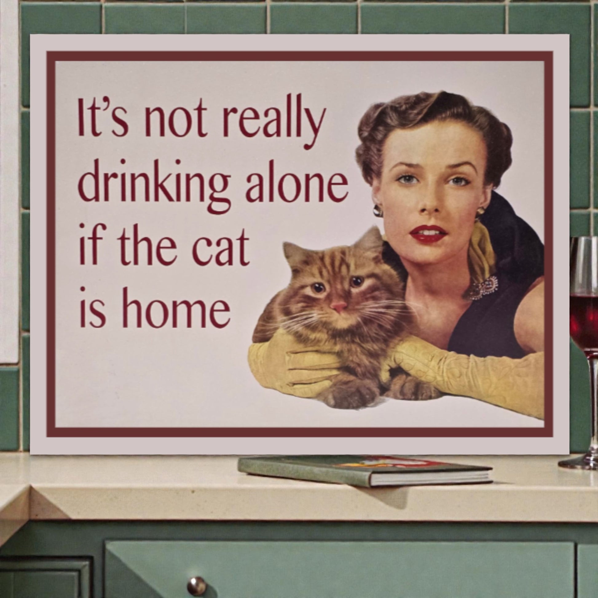 Poster of a woman holding a cat with text 'It's not really drinking alone if the cat is home' on a kitchen counter.