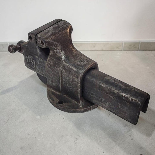 A vintage dark metal bench vise with a heavy-duty cast iron body, featuring a mounting base and a cylindrical work holder, photographed on a concrete floor against a white wall.