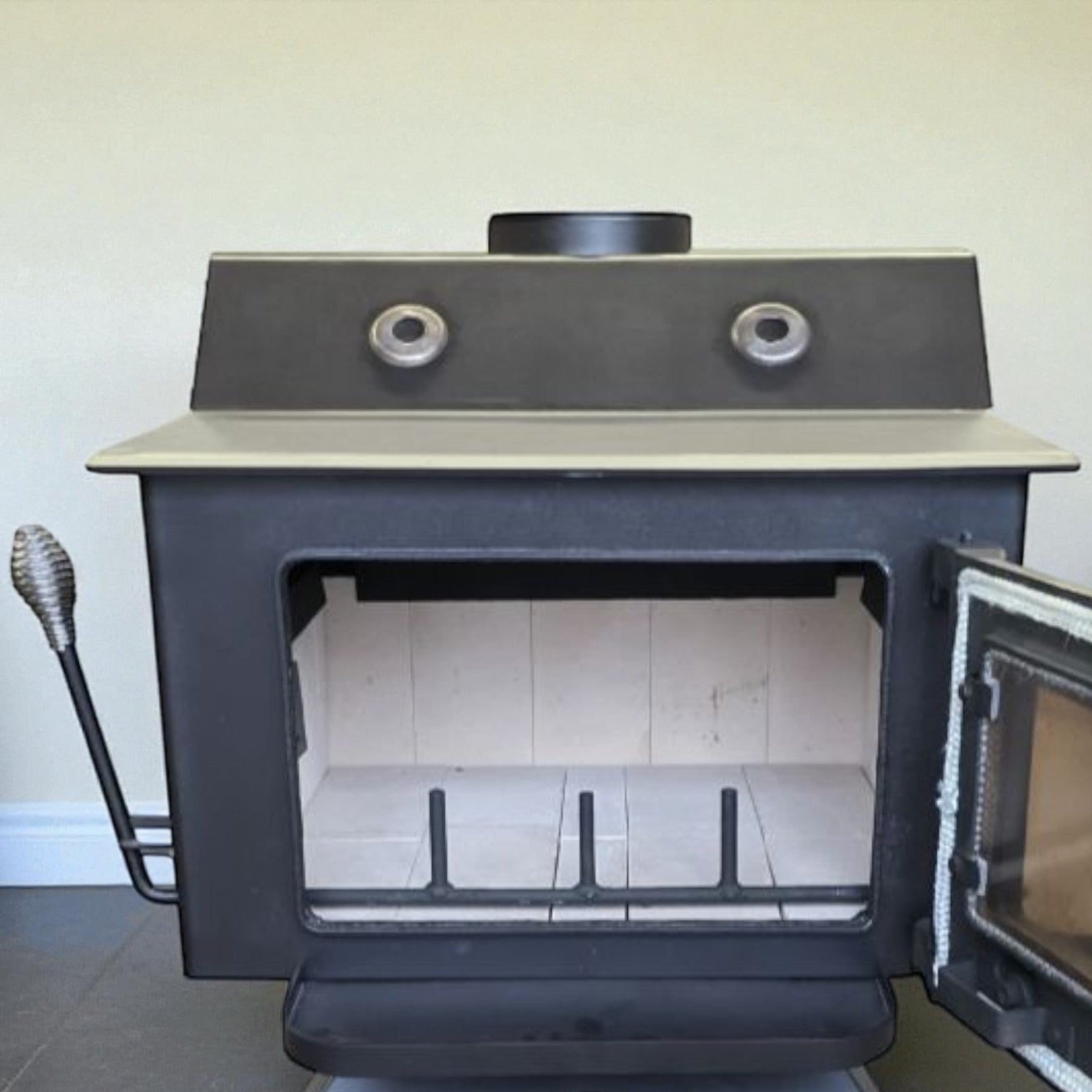 A black cast iron wood-burning stove with its door open, showing the interior firebox and a light-colored brick interior. The stove has a beige top panel, a silver support base, and a black handle with a textured grip on the left side.