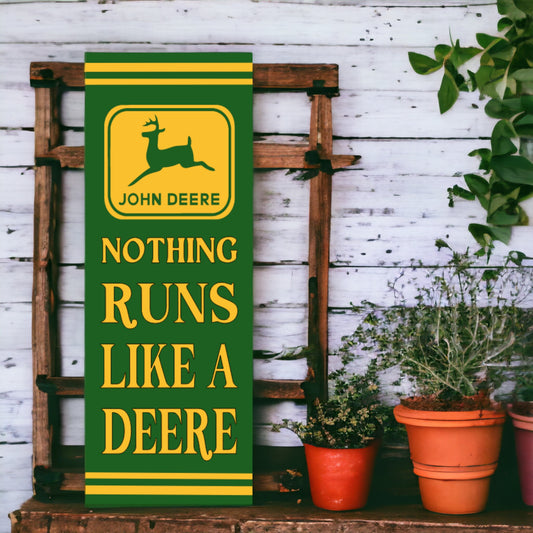 A vertical, retro style John Deere sign featuring the brand logo with the text 'NOTHING RUNS LIKE A DEERE' in bold lettering, placed against a wooden backdrop with green plants to the side.