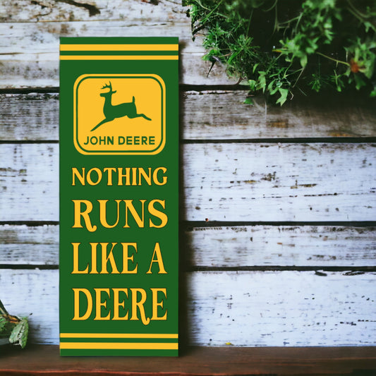 A vertical, retro style John Deere sign featuring the brand logo with the text 'NOTHING RUNS LIKE A DEERE' in bold lettering, placed against a wooden backdrop with green plants to the side.