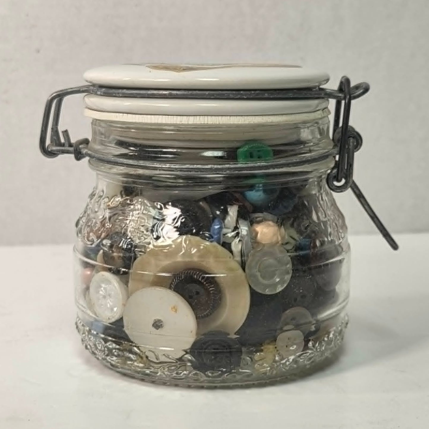 Clear glass jar with metal clasp lid filled with various buttons on a white background