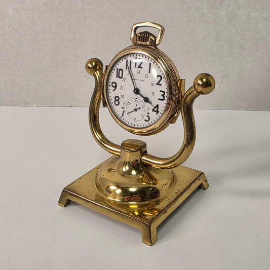 Gold pocket watch on a stand with a white face against a light gray background